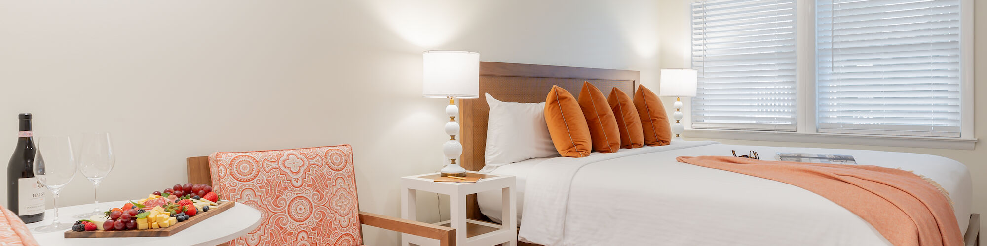 A cozy bedroom with a large bed, orange accents, a small table with two chairs, a wine bottle, and a fruit plate, under warm lighting.