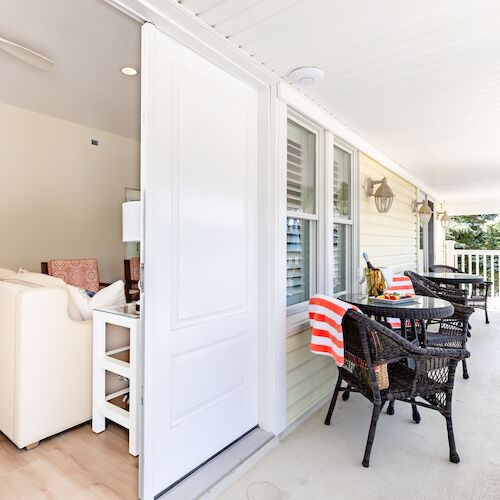 A cozy porch with wicker chairs and striped towels, overlooking a living room with light furniture through an open door.