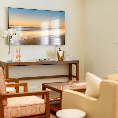 A cozy, modern living room with a wall-mounted TV, decorative items on a console table, comfy seating, and natural light coming through blinds.