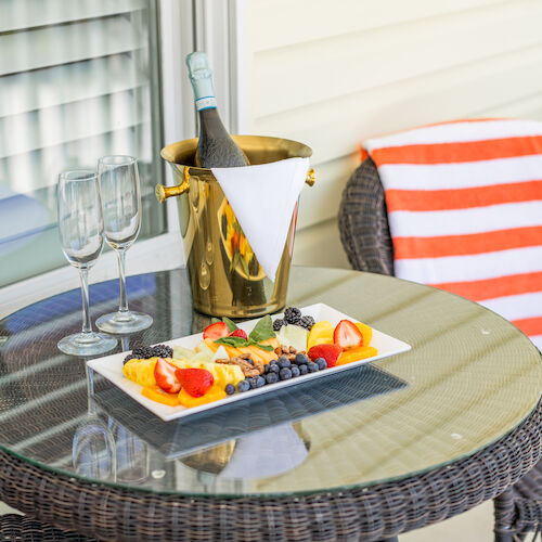 A glass table with a fruit plate, two empty glasses, and a champagne bottle in a golden ice bucket, with a chair draped with a striped towel.