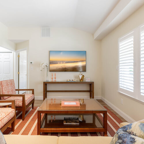 A cozy living room with a TV, orange-patterned chairs, coffee table, and a striped rug, bathed in natural light from large windows with blinds.