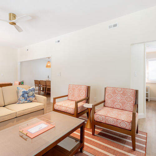 This image shows a modern living room with a beige couch, two patterned armchairs, coffee table, chandelier, and an adjacent dining area.