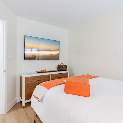 A neatly arranged bedroom with a TV mounted on the wall, an orange-accented bed, a wooden stand, and a glimpse into an adjacent room.