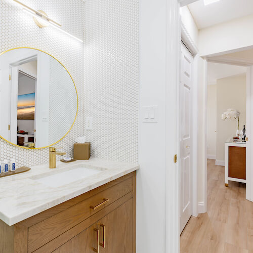 A modern bathroom with a round gold-framed mirror above a wooden vanity, next to the entrance of a hallway and another room featuring a table with flowers.