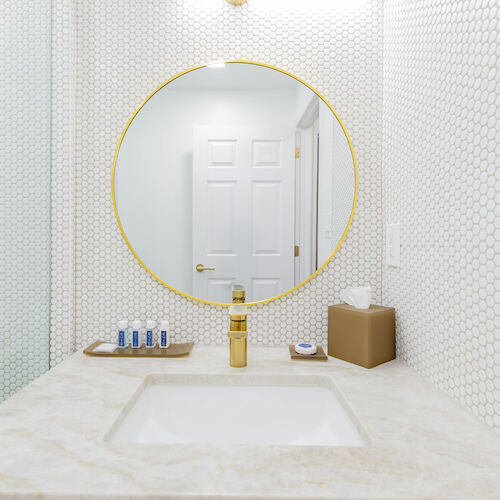 This image shows a bathroom vanity with a round mirror, modern sink, toiletries, a brown tissue box, and a door in the background.