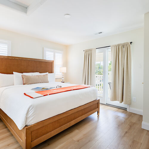 A bright bedroom with a large bed, bedside tables, lamps, a dresser with flowers, and curtains leading to a balcony.