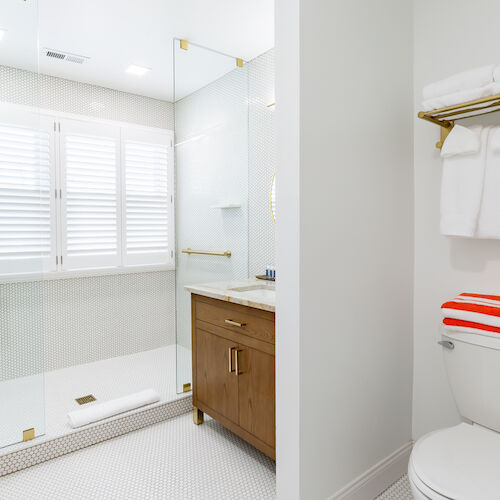 The image shows a clean, modern bathroom with a glass shower, wooden vanity, white towels on a shelf, and a toilet with a red-striped towel.