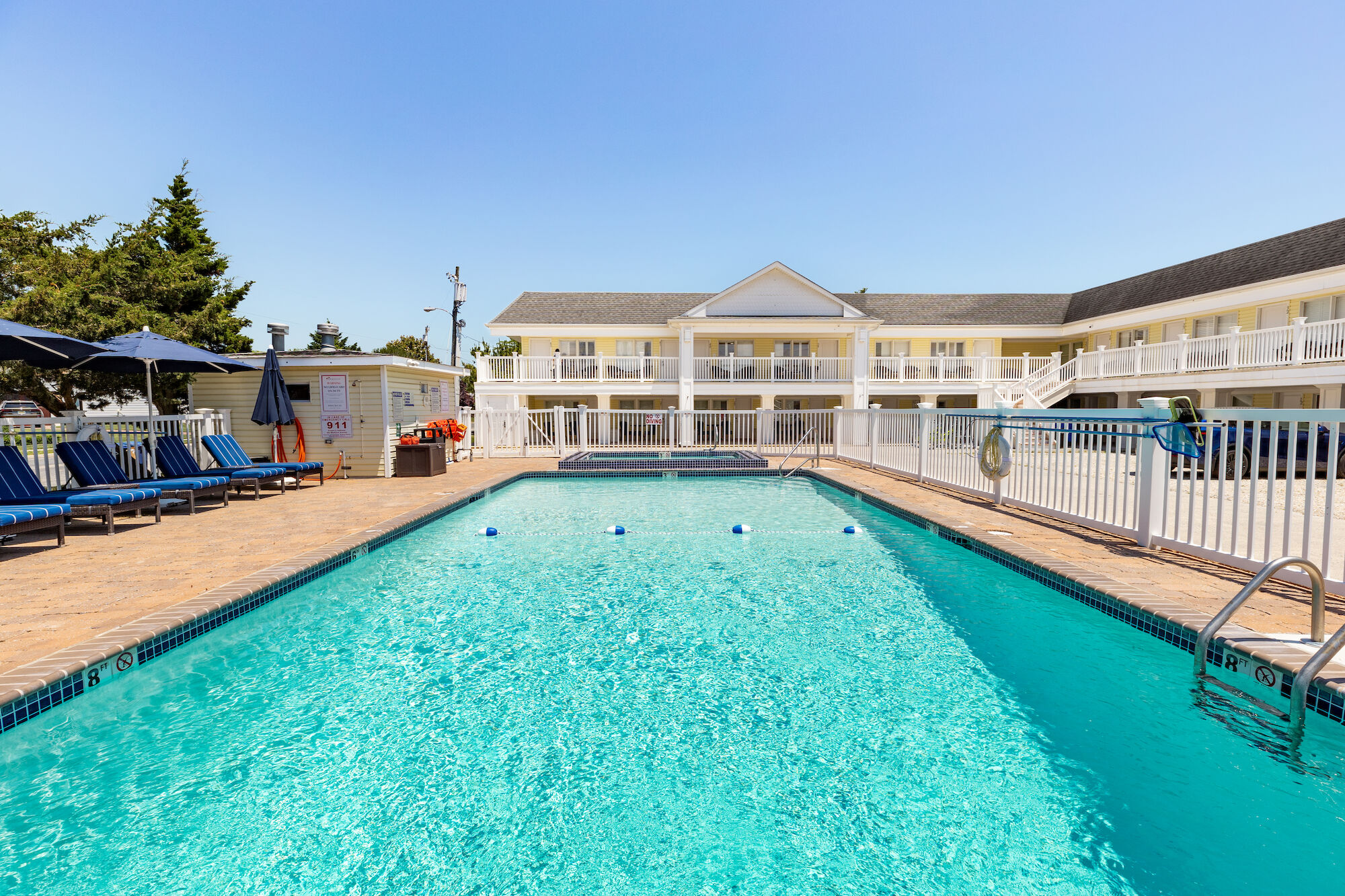 The image shows an outdoor swimming pool surrounded by lounge chairs and umbrellas, with a two-story white building in the background.