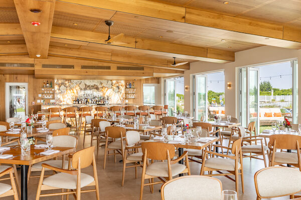 This image shows a modern, well-lit restaurant with wooden beams, tables set for dining, and large windows leading to an outdoor seating area.