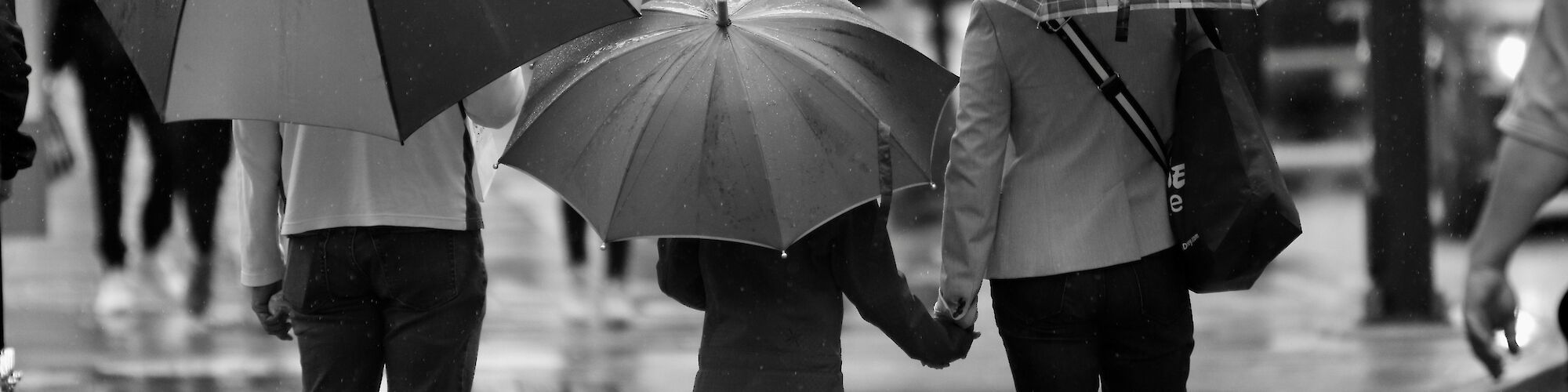 A black and white photo showing three people walking in the rain with umbrellas, holding hands in a cityscape setting.