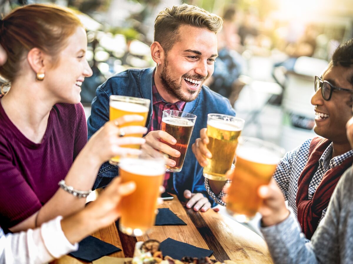 A group of friends sits around a table, smiling and raising their glasses filled with beer, enjoying their time together at a social gathering.