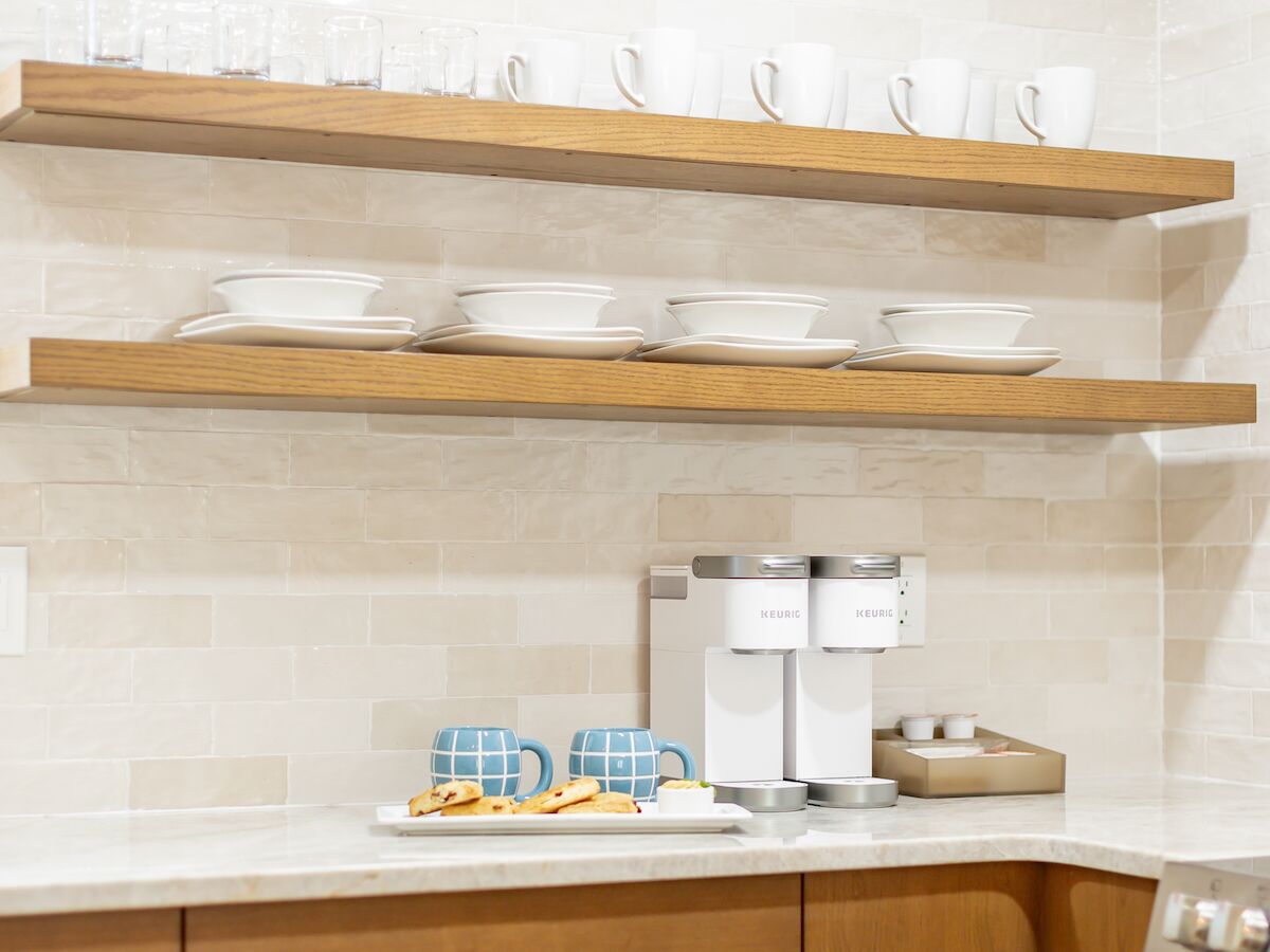 The image shows a kitchen setup with two wooden shelves holding white cups and plates, a coffee machine, and checkered mugs on the countertop.