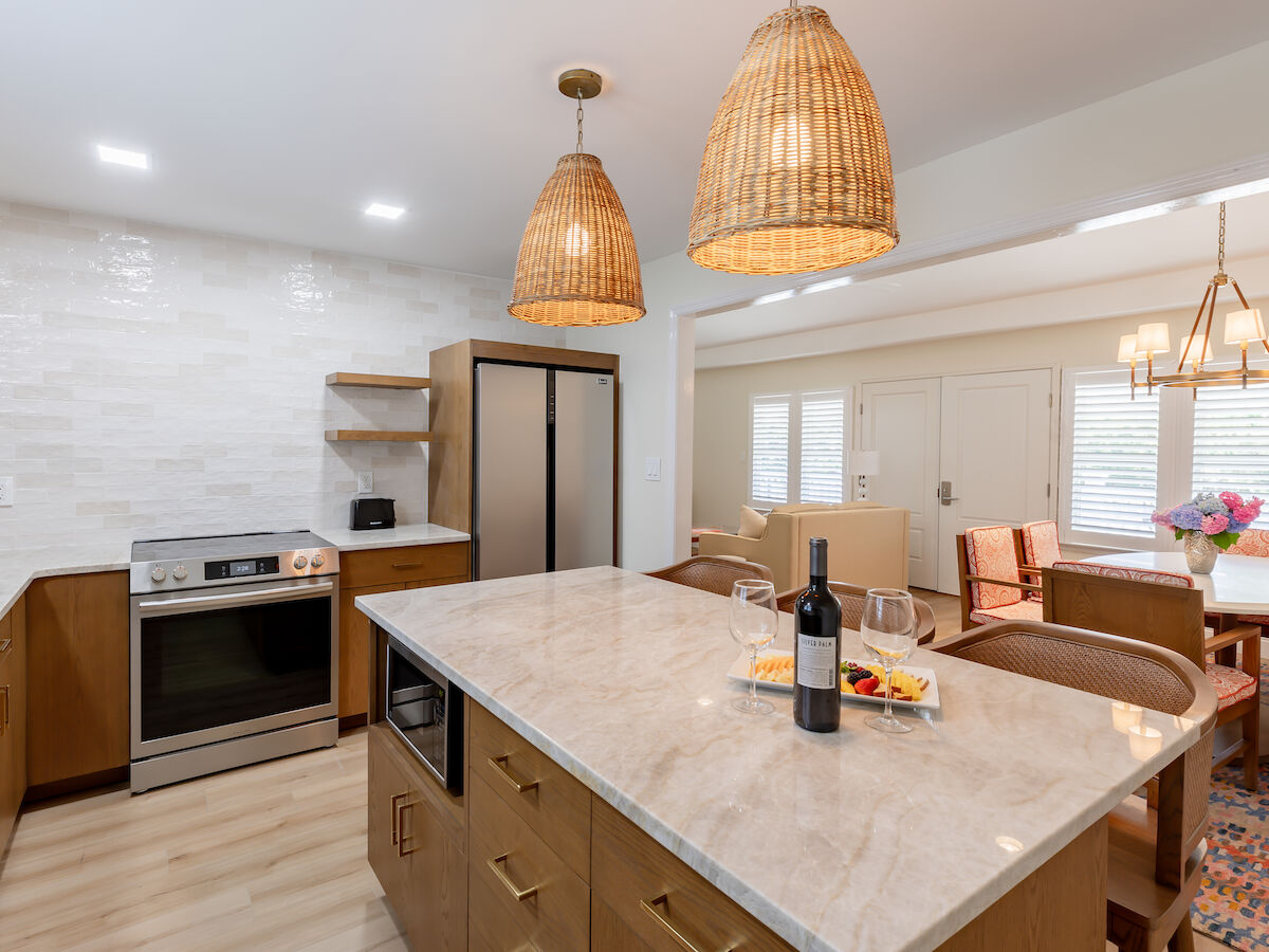 A modern kitchen with wooden cabinets, an island with a wine bottle and cheese platter, wicker pendant lights, an oven, fridge, and dining area.