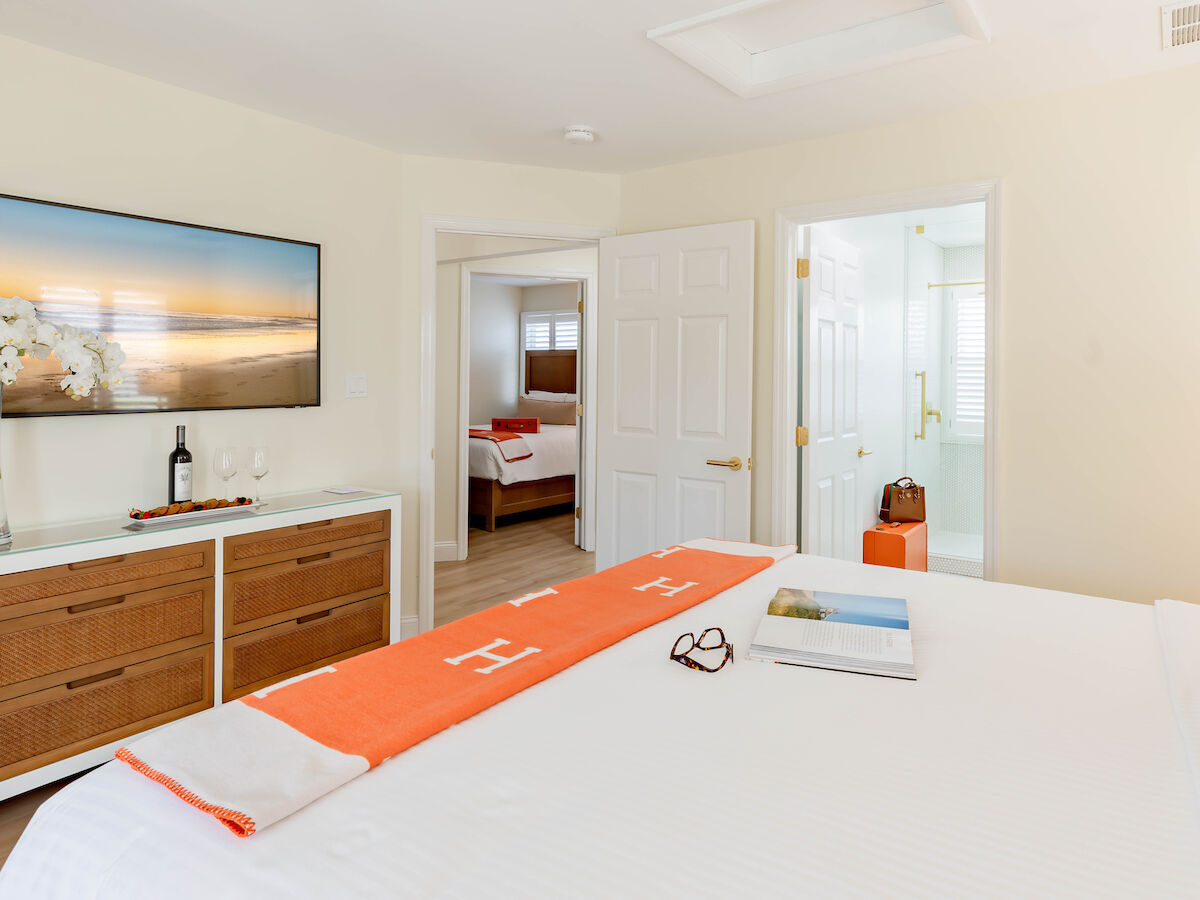 A bedroom with a bed, a dresser, a TV displaying a beach scene, a tray with glasses, a book, and a pair of glasses. Adjacent room and bathroom are visible.