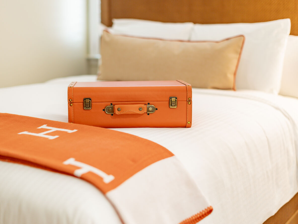 An orange suitcase is placed on a neatly made bed with white bedding and a matching orange blanket featuring the letter 