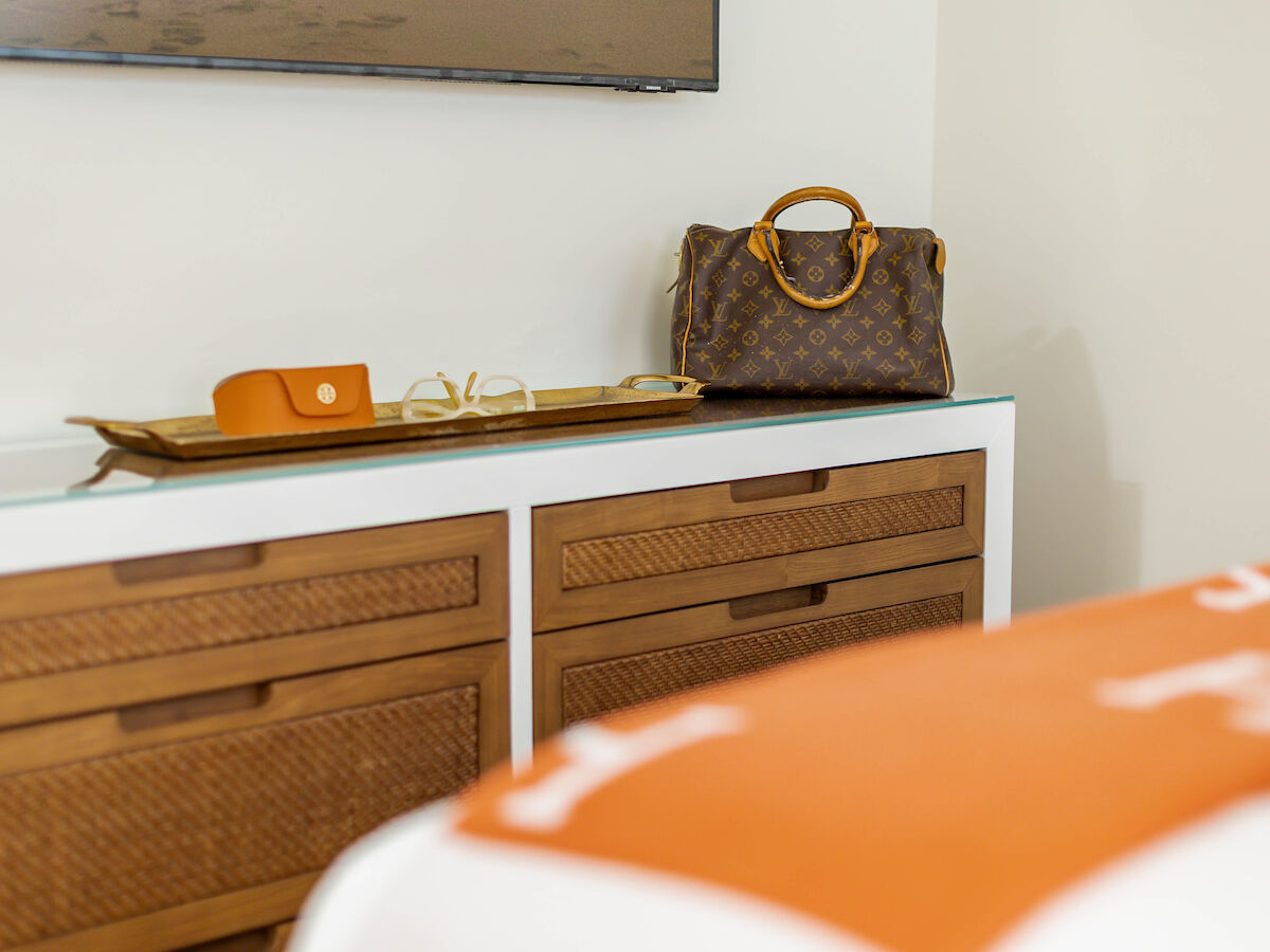 A dresser topped with a designer handbag, glasses on a tray, and a small orange item in a room with a bed.