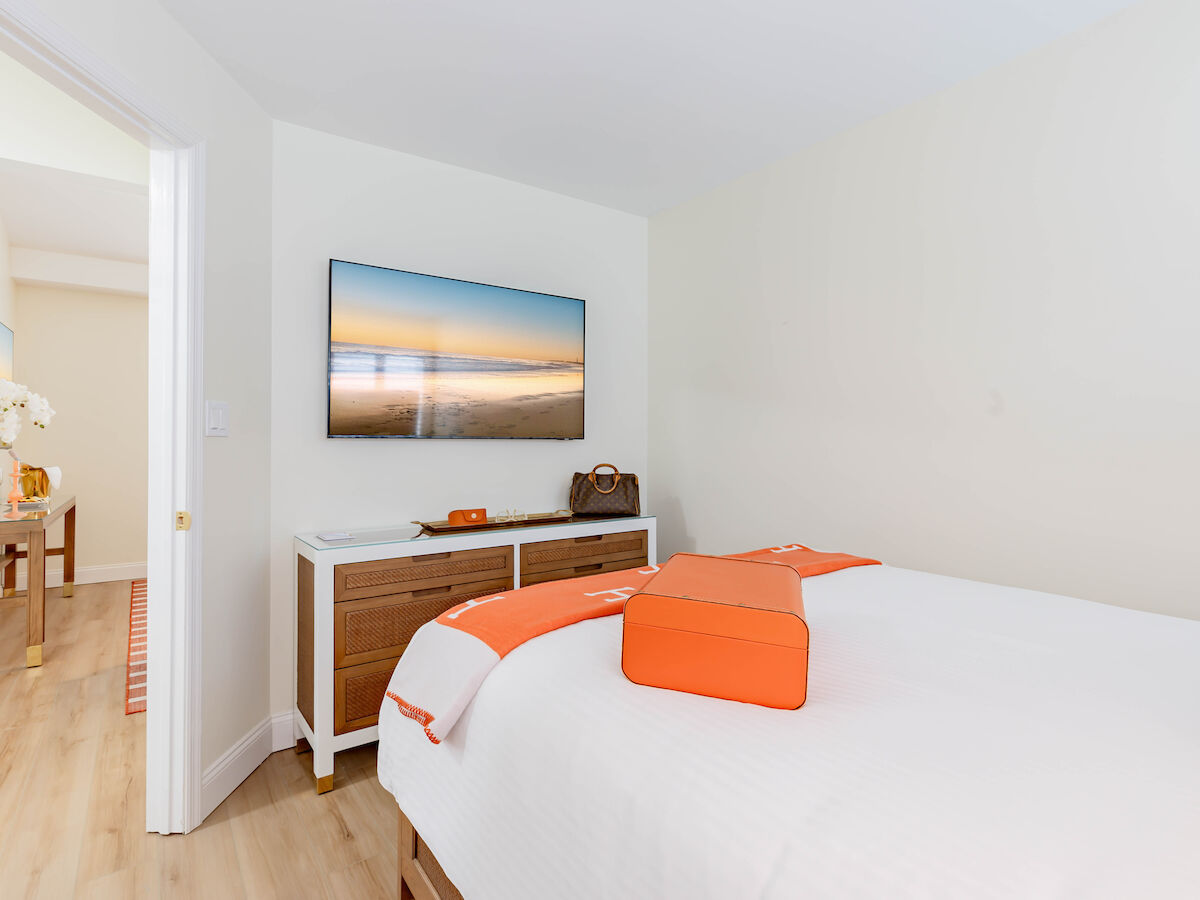 A bright bedroom with a neatly made bed with orange accents, a TV on the wall, a dresser, and an open door leading to another room.