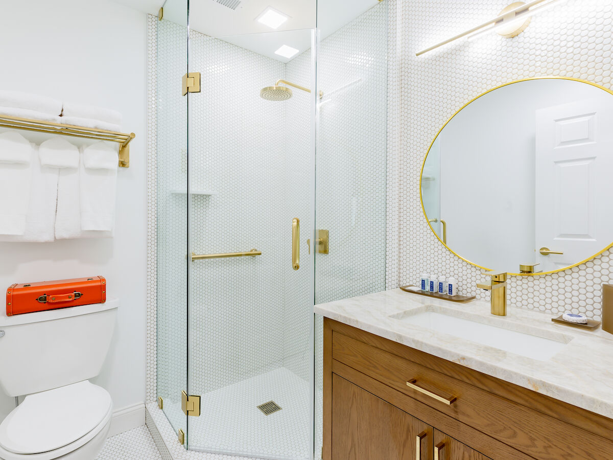 A modern bathroom with a glass shower, a toilet, a wooden vanity with a round mirror, towels on a rack, and toiletries on the countertop.