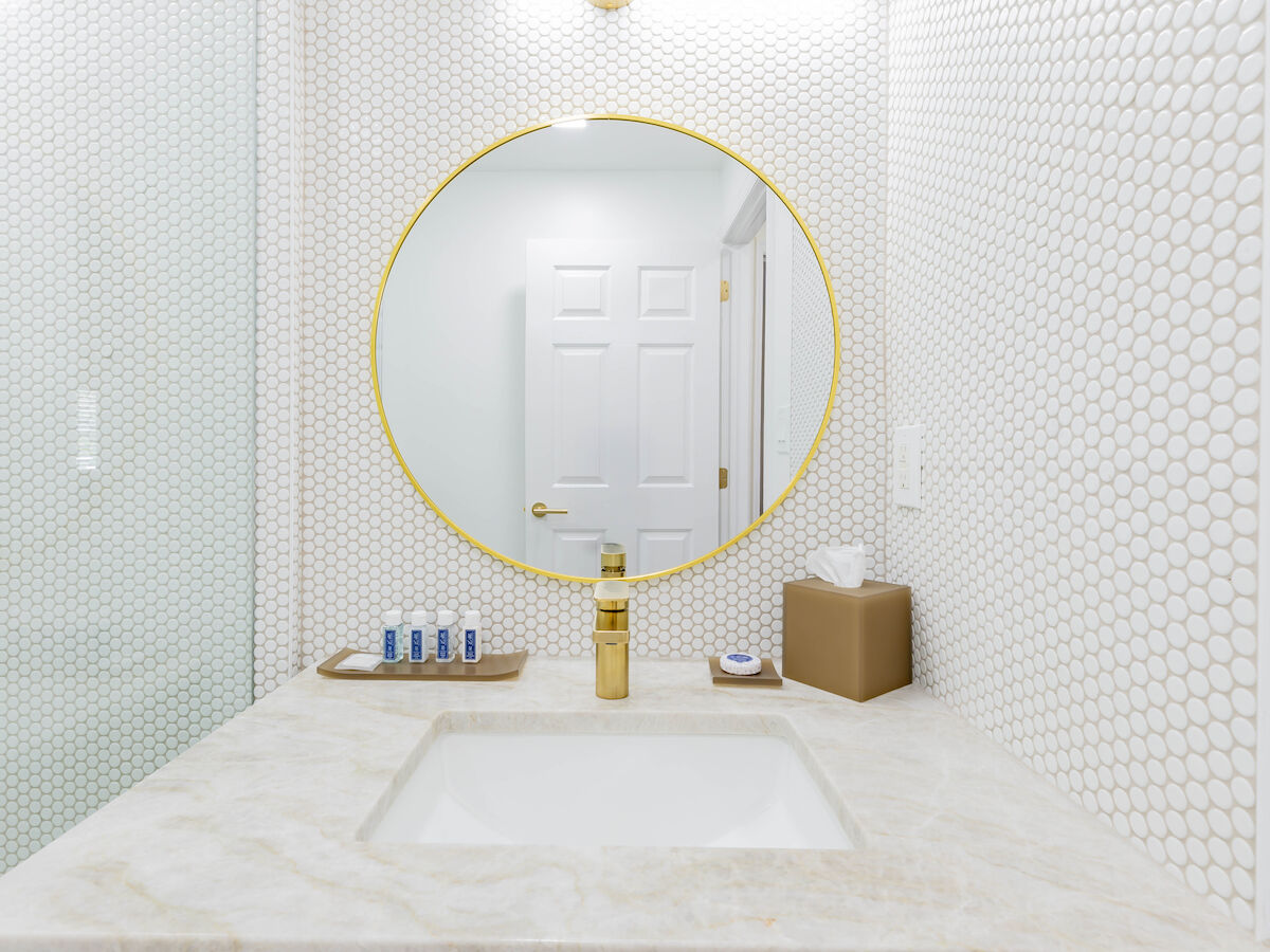 A bathroom with a white countertop, a golden faucet, a round mirror, brown tissue box, toiletries, and white hexagonal tile walls is shown.