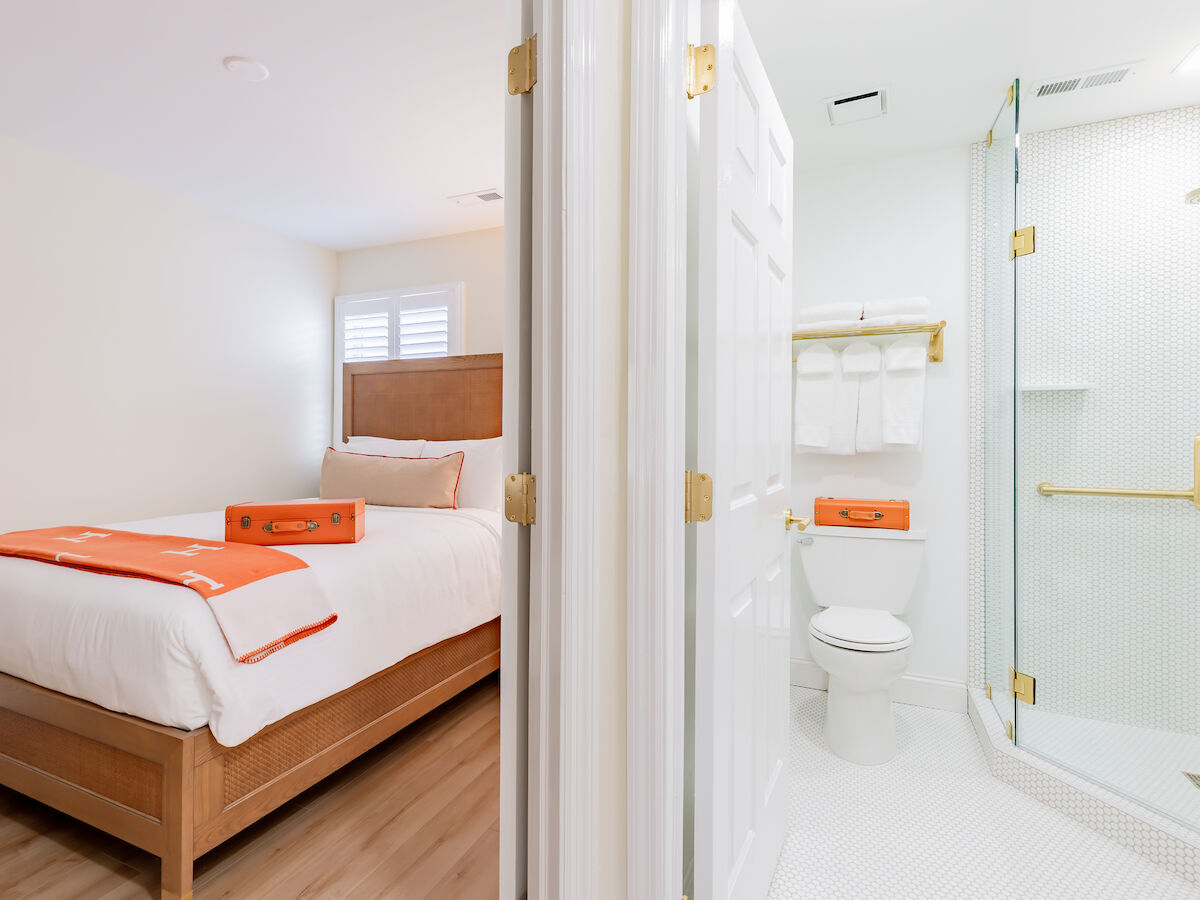 A bright, tidy bedroom and bathroom with a shower, toilet, bed, and orange accents on white linens and towels, ending the sentence.