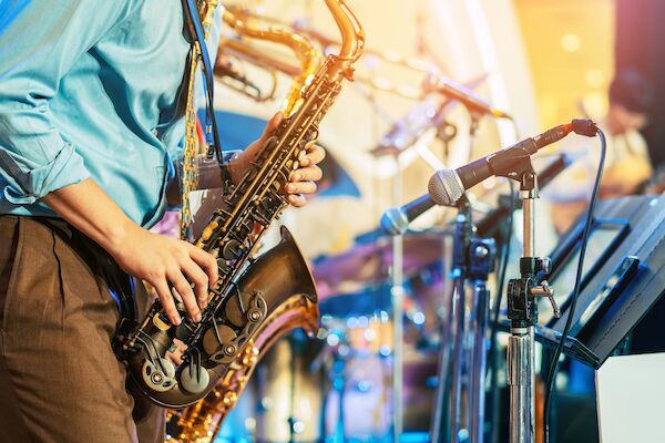 A musician playing a saxophone on stage, with drums and other equipment in the background, indicating a live performance setting.