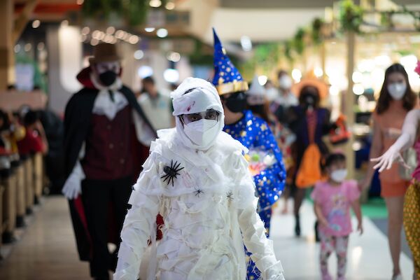 People dressed in various costumes, including a mummy, wizard, and vampire, walking indoors while wearing masks, possibly for a festive event.