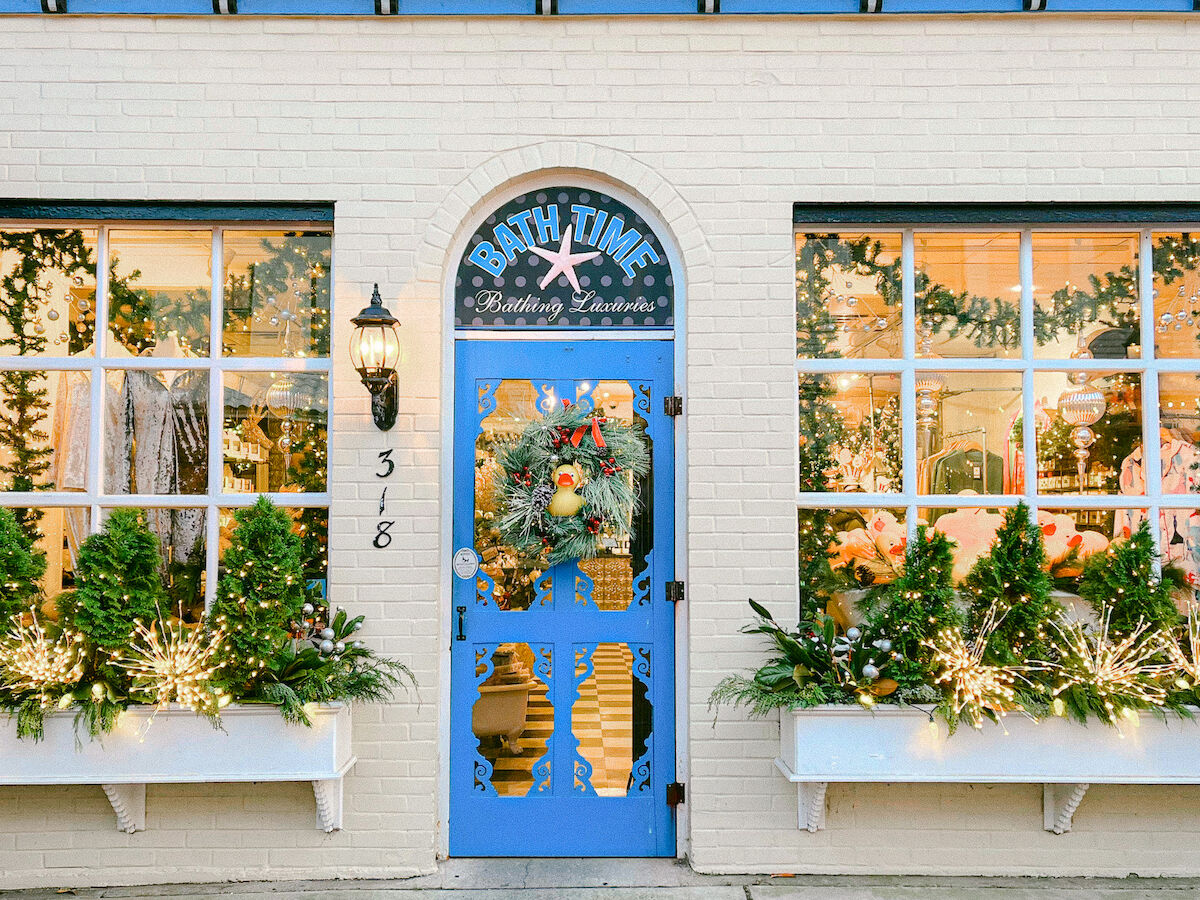 Storefront of &ldquo;Bath Time&mdash;Bathing Luxuries&rdquo; with a blue door, Christmas lights, garlands, and a festive wreath in the window.