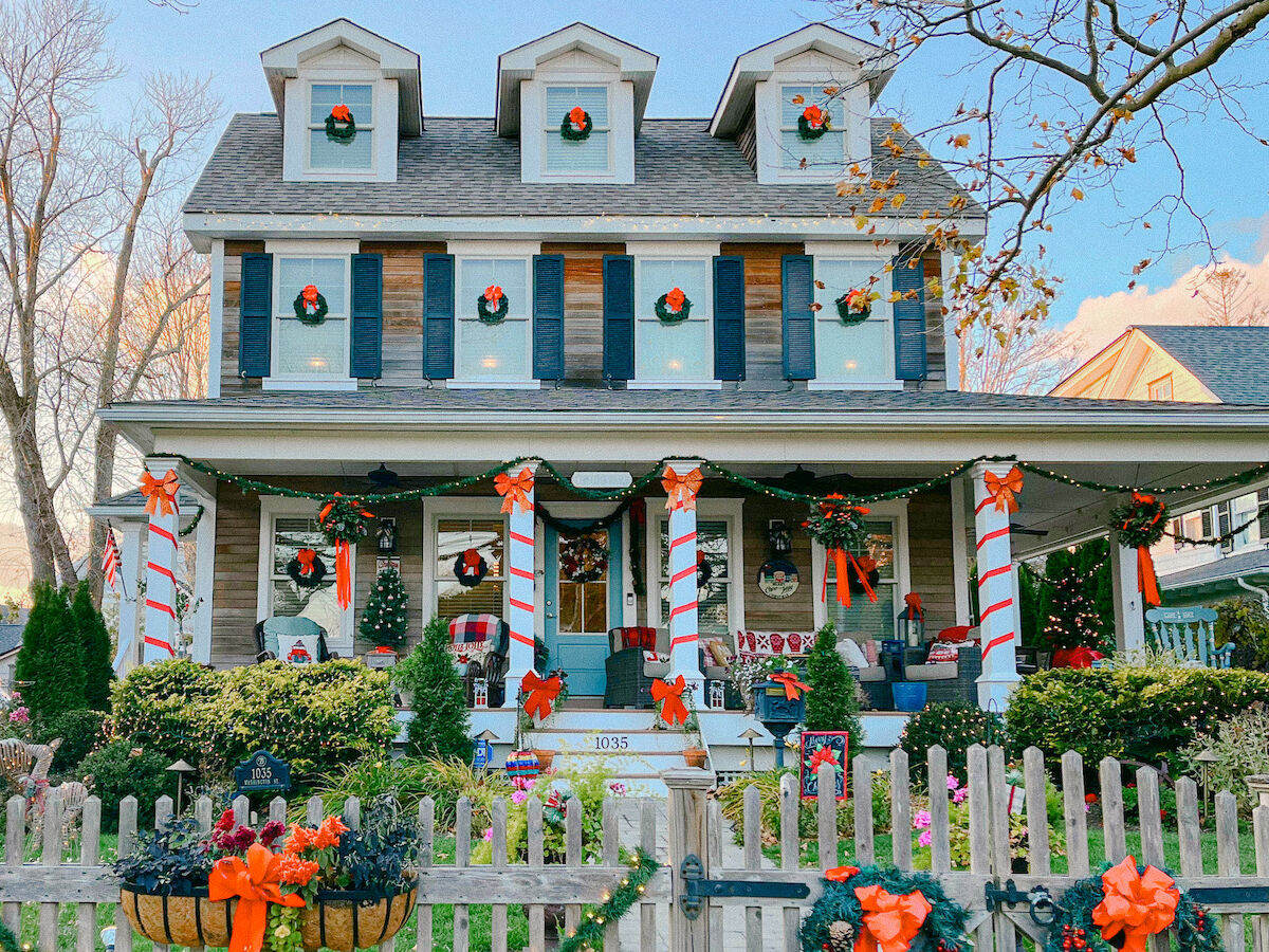 A Christmas-decorated house with wreaths, candy-cane yard banners, string lights, and seasonal decorations in front of a fence.