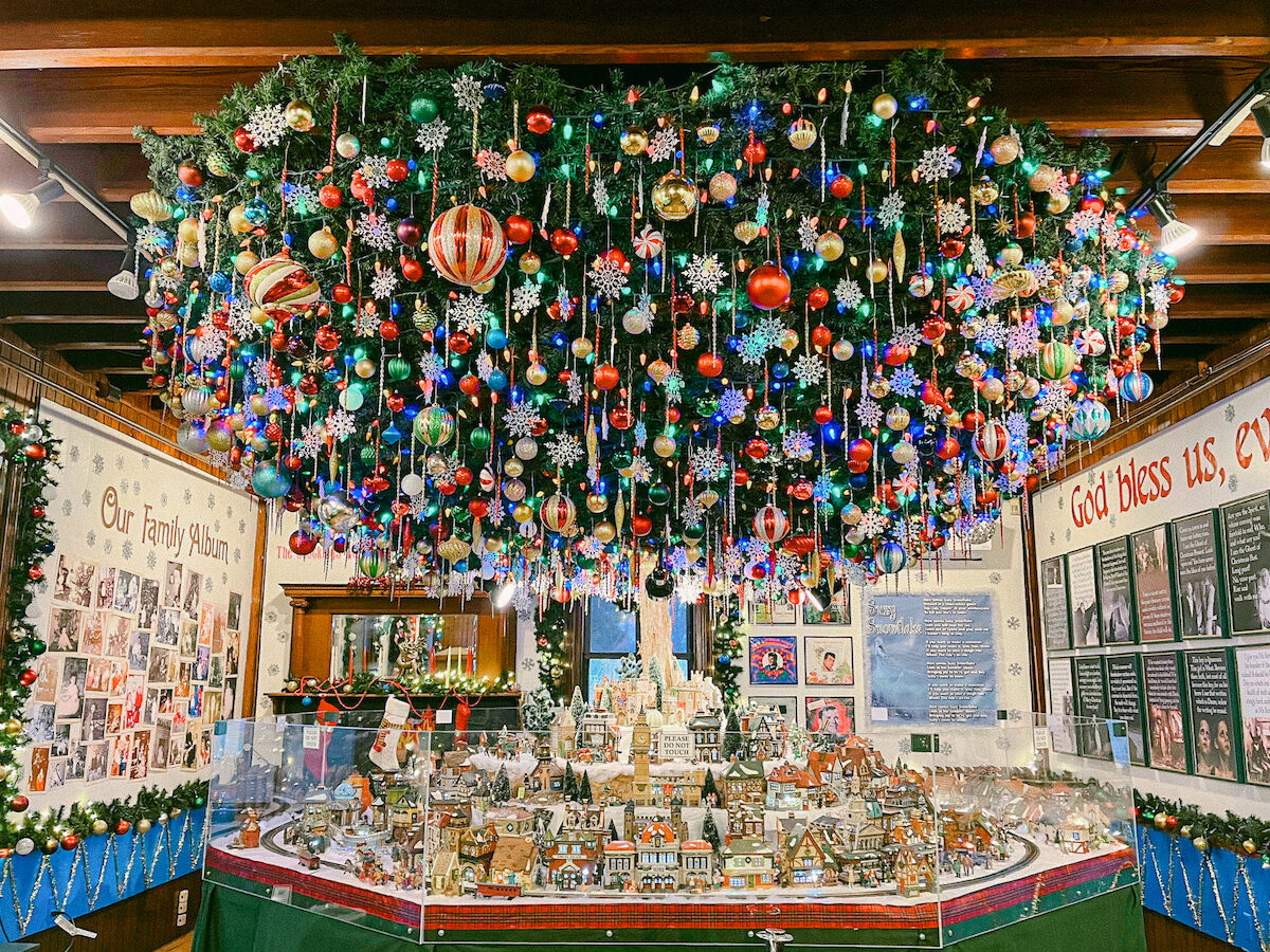A towering Christmas tree packed with colorful ornaments and snowflakes hangs over a winter village display of houses.