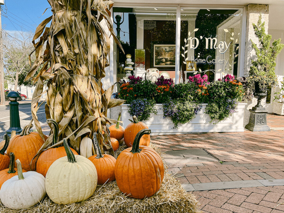 A storefront decorated with pumpkins, corn stalks, and flowers is pictured with a hay bale on a brick pavement.