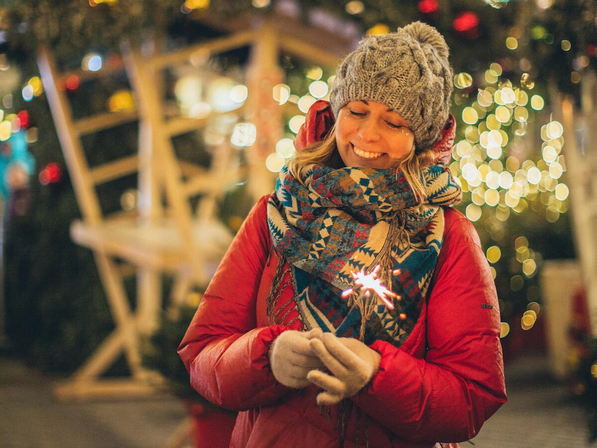 A smiling person in a red winter coat and knitted hat stands in the snow, surrounded by glowing holiday lights and sparkly bokeh.