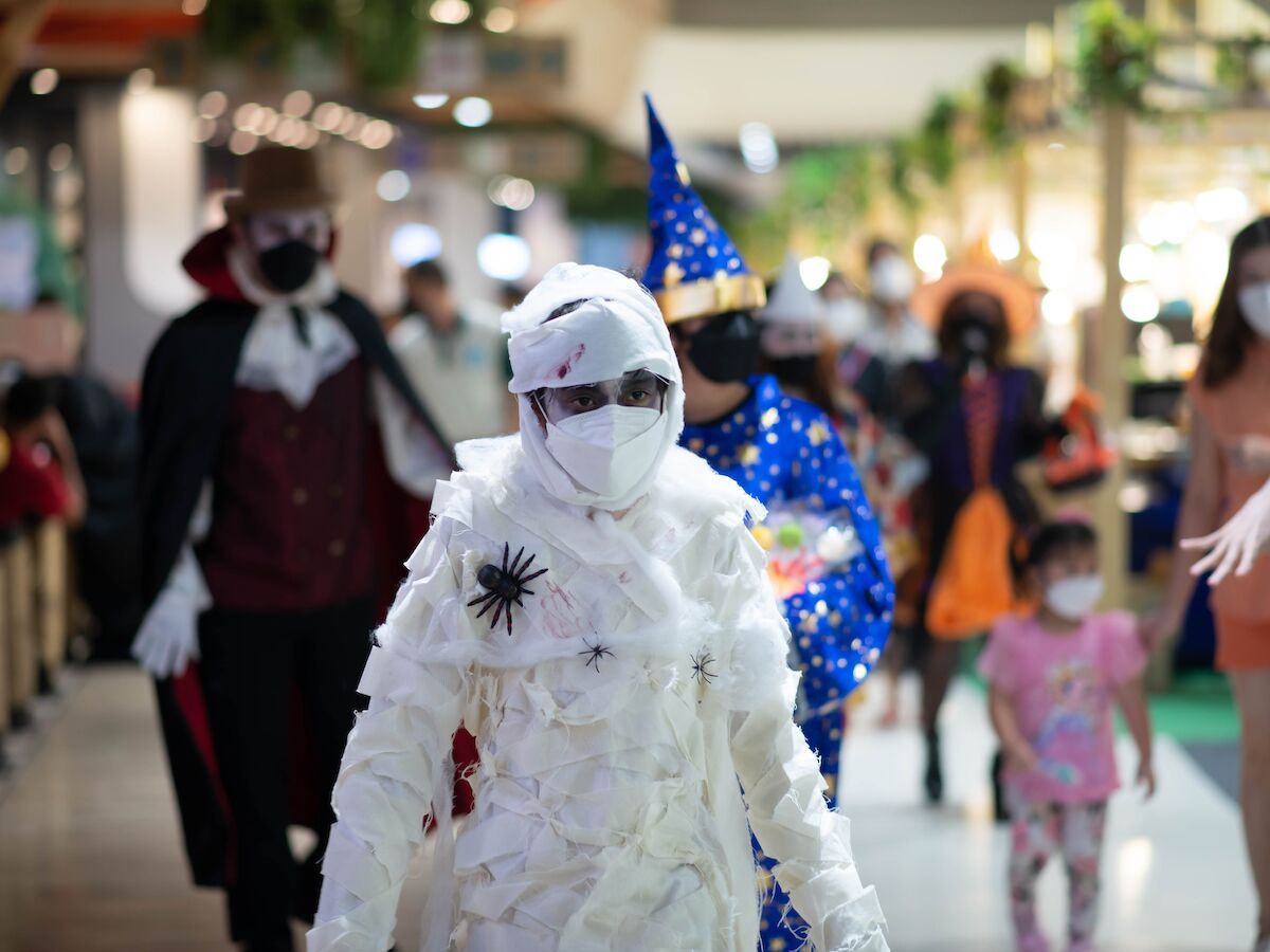 People dressed in various costumes, including a mummy, magician, and others, are walking in what seems to be a Halloween or costume event.