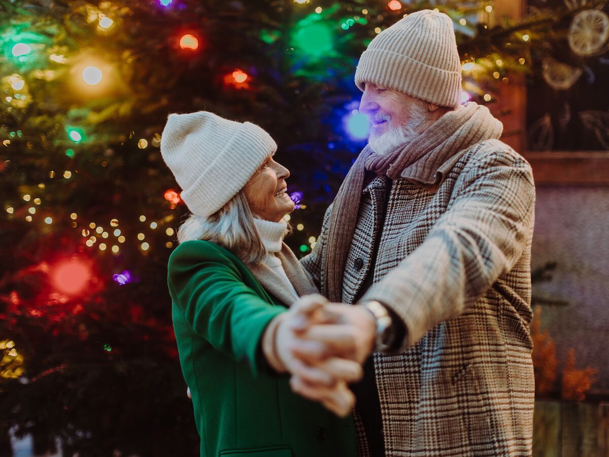 An elderly couple dances joyfully in front of a brightly lit Christmas tree, wearing warm winter clothing.