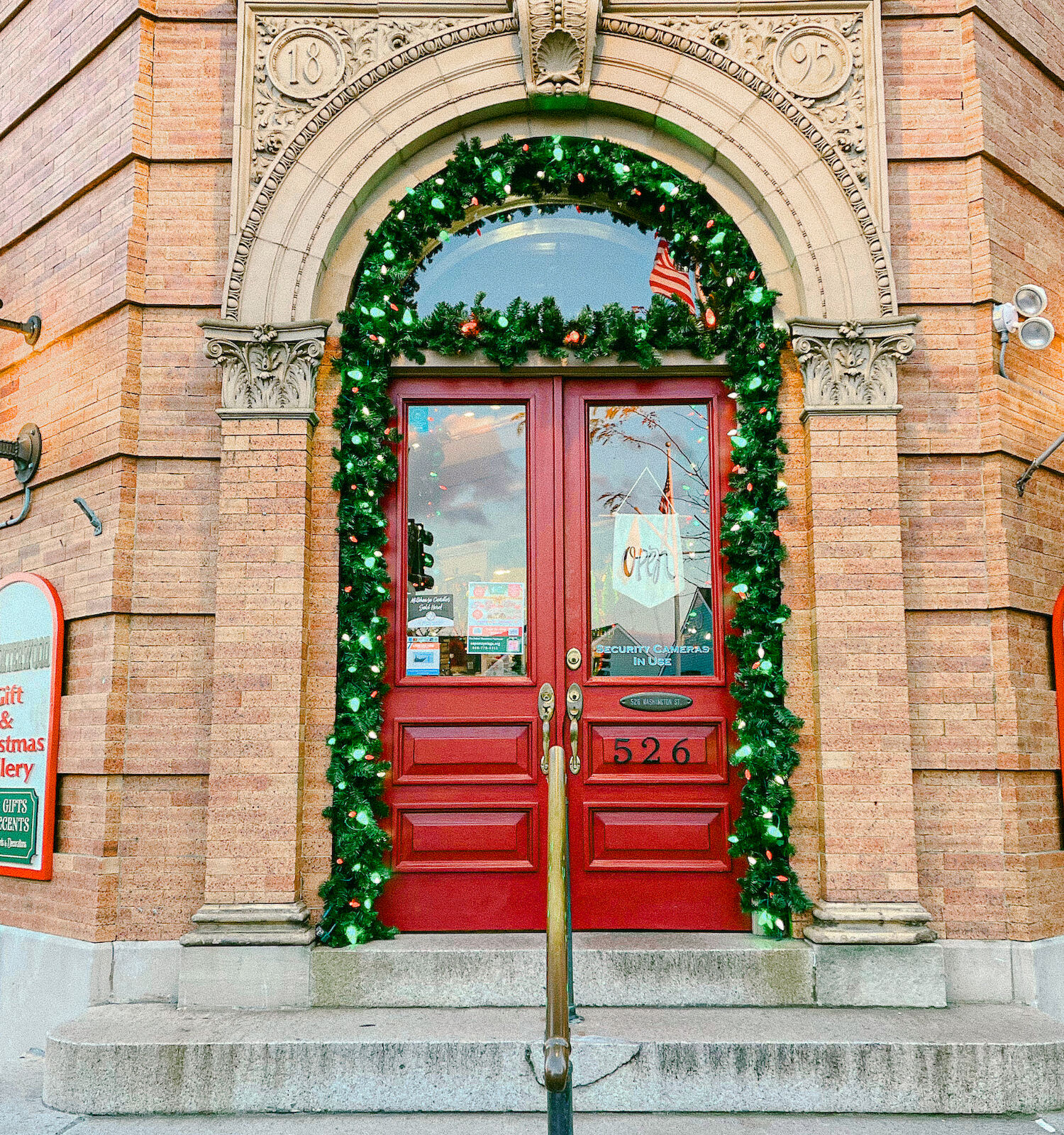 A storefront with red doors, surrounded by festive garland. Signs indicate it's a Christmas shop. Holiday decorations are visible inside.