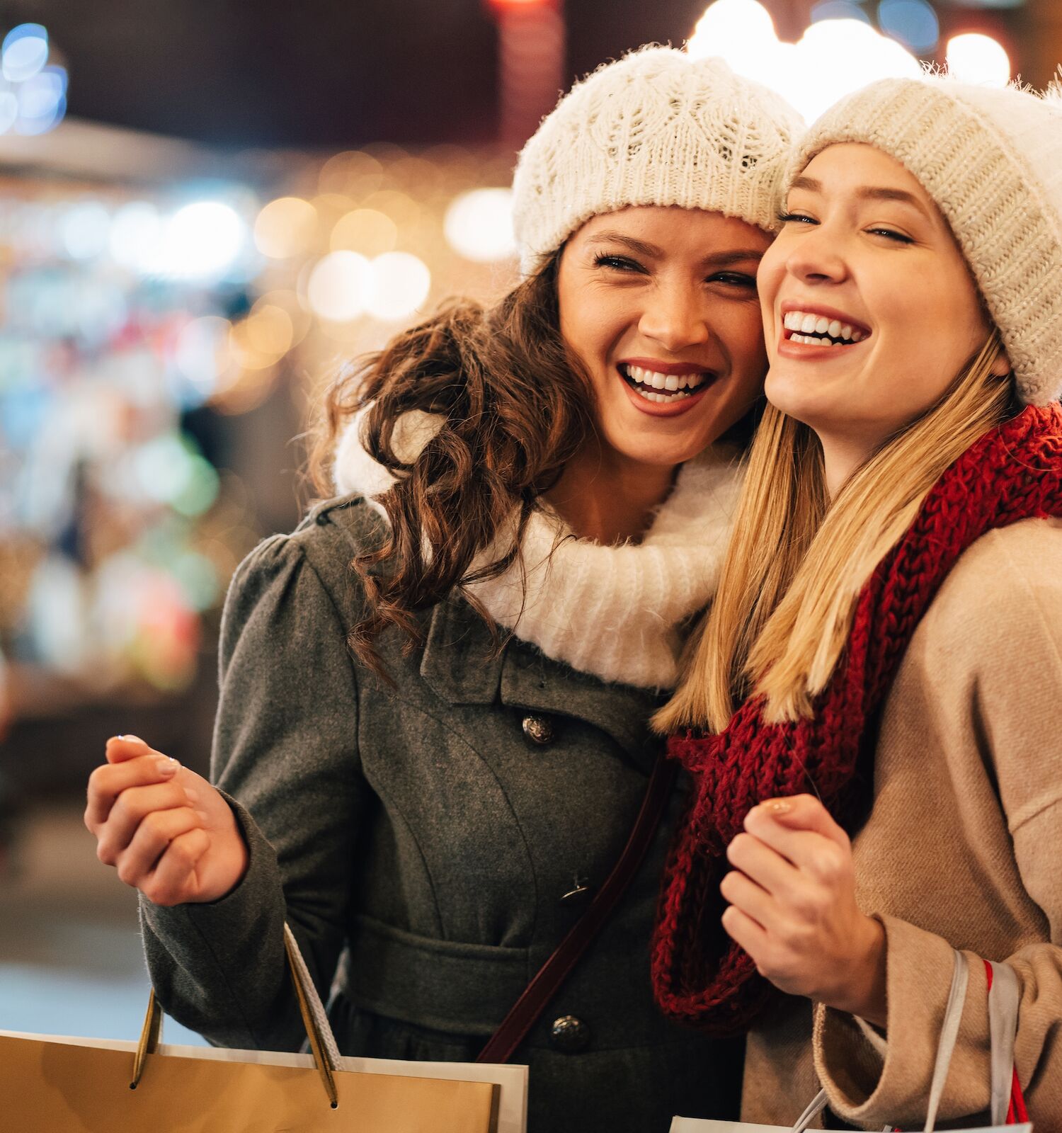 Two people are smiling and holding shopping bags outdoors, dressed warmly with hats and scarves, in a festive setting at night.