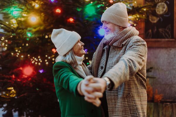 An older couple in winter clothing embraces in front of a Christmas tree with colorful lights, smiling at each other.