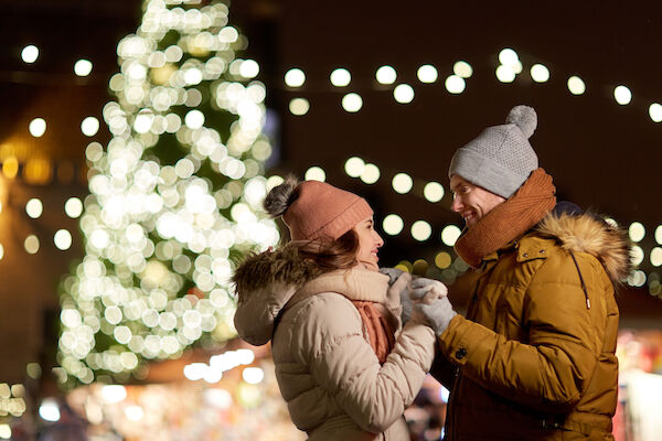 A couple in winter clothing enjoys a festive atmosphere, with a beautifully lit Christmas tree and string lights in the background.