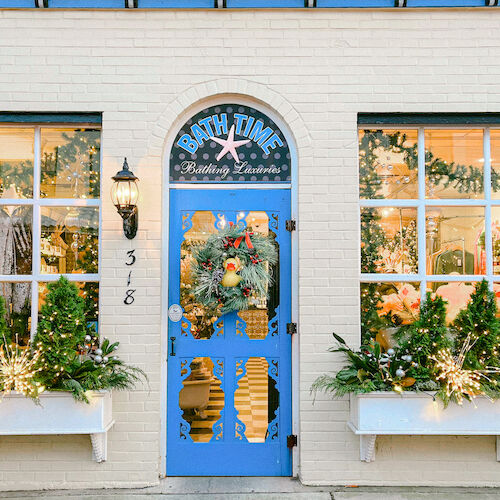 A festive storefront with a blue door, holiday wreath, and window displays featuring greenery and decorations, creating a cheerful atmosphere.