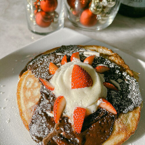 A pancake topped with chocolate, cream, and strawberries. Decorative ornaments and a bottle in the background complete the scene.