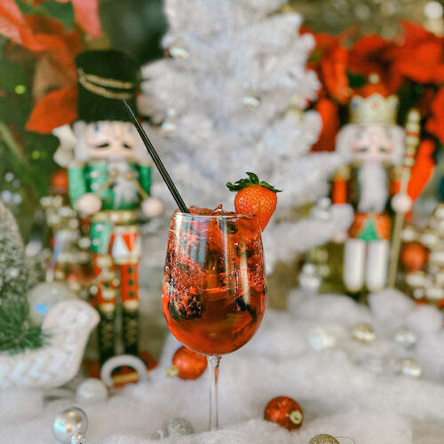 A festive scene with a decorated Christmas tree, nutcracker figures, and a strawberry cocktail surrounded by ornaments and faux snow.