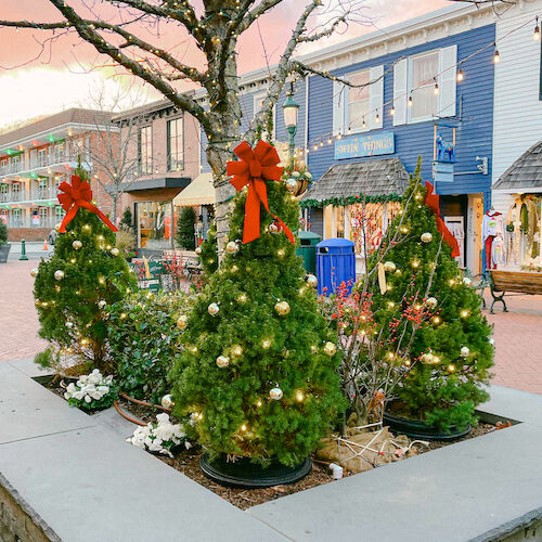 A festive outdoor scene features Christmas trees with red bows and ornaments, near shops and benches adorned with holiday decorations.