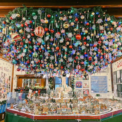 A festive room with a large, colorful Christmas wreath suspended from the ceiling and a detailed holiday village display underneath.