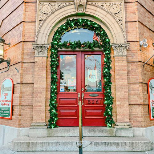 A storefront with red doors is decorated with holiday garlands. Signs indicate it's a gift and Christmas gallery.