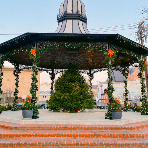 A decorated gazebo with garlands and a Christmas tree inside, set in a park with brick steps and some trees around.