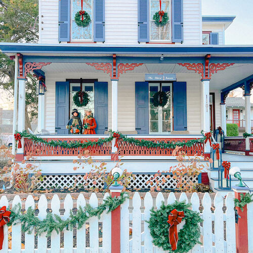 A festive house with blue shutters, porch decorations, wreaths, and garlands on a picket fence, exuding a holiday spirit.