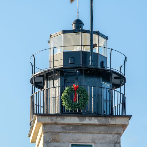 A lighthouse lantern room with a weather vane on top and a festive wreath hung on its exterior railing against a clear blue sky.