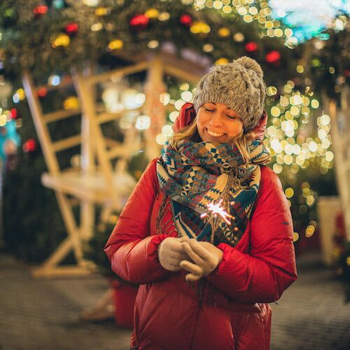 A person in winter clothing stands in a festive setting with lights and decorations, possibly at a holiday market or celebration.