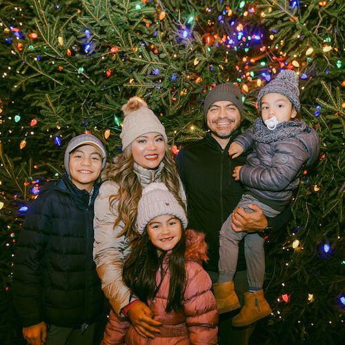 A family posing happily in front of a Christmas tree adorned with colorful lights, wearing warm winter clothing and smiling.