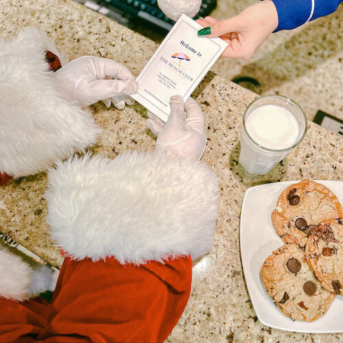 A person in a red suit receives a card. Nearby, a plate of cookies and a glass of milk are on the counter.