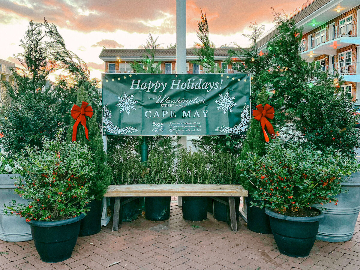 Holiday display with a “Happy Holidays! Washington Street, Cape May” sign, decorated with festive greenery and red bows, near a bench.